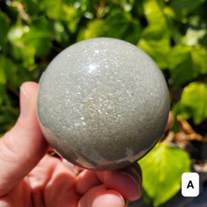 May include: A smooth, gray sphere with a speckled surface is held in a hand. The sphere has a glossy finish and reflects light. The background is blurred green foliage. The sphere is a decorative item.