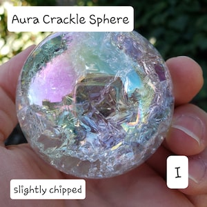 May include: Aura Crackle Sphere with iridescent colors. The clear sphere has a cracked texture and is slightly chipped. The text "Aura Crackle Sphere" is at the top, and "slightly chipped" is at the bottom.