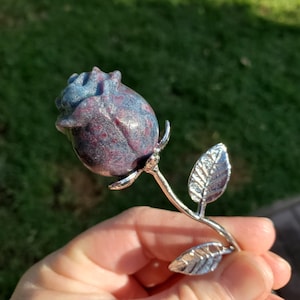 May include: A carved stone rose bud in shades of blue and red, mounted on a silver-colored metal stem with two leaves. The rose is held in a hand against a blurred green background. The rose is a decorative item.