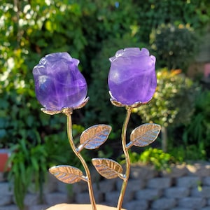 May include: Two amethyst rose sculptures with gold-colored stems and leaves. The purple rosebuds are carved from translucent stone. The leaves have a detailed, textured appearance. The background is blurred, with green foliage.