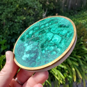 Può includere: Un sottobicchiere rotondo in malachite verde con un bordo dorato. Il sottobicchiere è realizzato in pietra naturale e presenta un motivo unico e vorticoso.