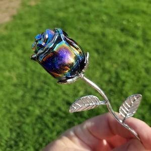 May include: A handcrafted iridescent rose with a metallic finish. The rose has a vibrant color gradient of blues, greens, and purples. The stem and leaves are silver-toned, and the rose is held against a blurred green background.