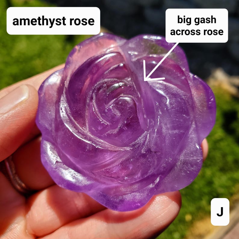 May include: A carved amethyst rose with a deep purple hue. The gemstone is shaped into a rose with detailed petals. The text "amethyst rose" is in a white box above the rose. A large gash is visible across the rose.