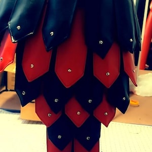 May include: A handmade, black and red leather garment, possibly armor or a skirt, displayed on a mannequin. The design features overlapping, scale-like pieces with silver rivets. The waist band is red with decorative stitching.