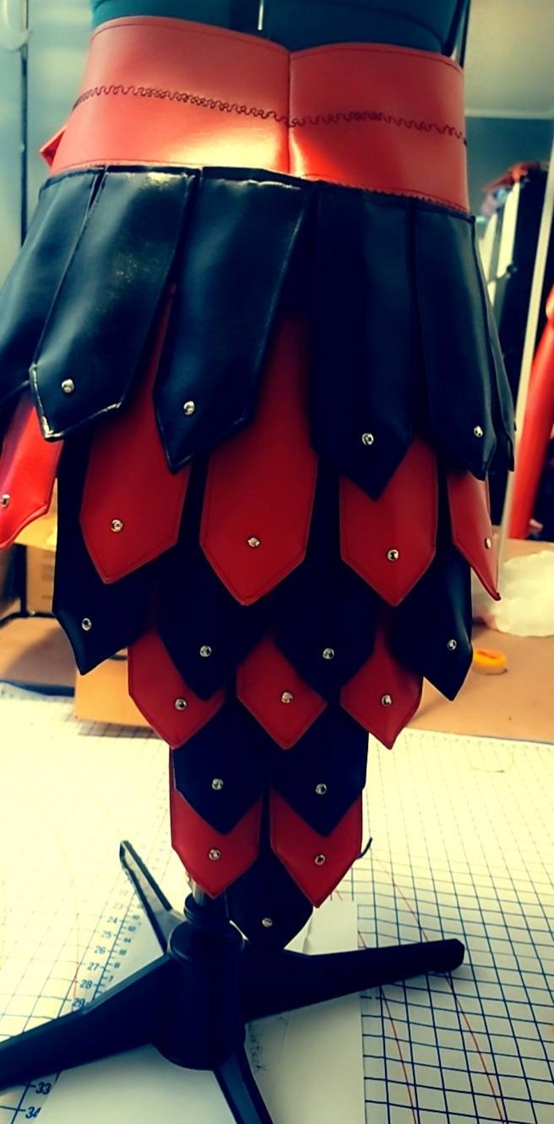 May include: A handmade, black and red leather garment, possibly armor or a skirt, displayed on a mannequin. The design features overlapping, scale-like pieces with silver rivets. The waist band is red with decorative stitching.