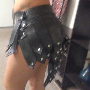 May include: Black leather skirt with layered, riveted panels. The skirt has a high waist and a unique, asymmetrical design. The skirt is a bold fashion statement.