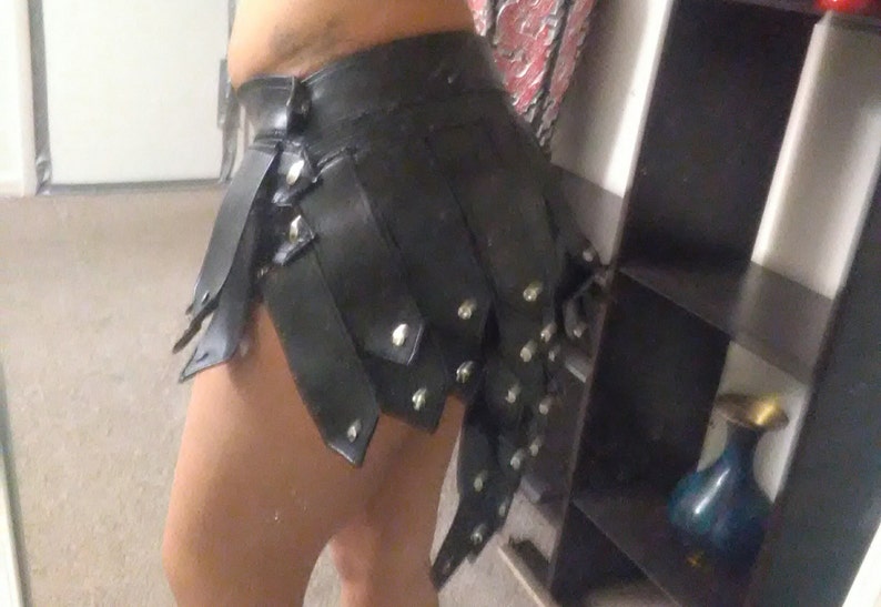 May include: Black leather skirt with layered, riveted panels. The skirt has a high waist and a unique, asymmetrical design. The skirt is a bold fashion statement.