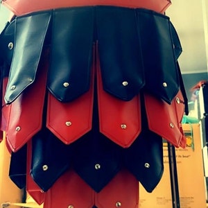 May include: A red and black leather skirt with a layered design. The skirt features alternating red and black panels, each secured with small silver rivets. The waist band is red and the skirt is displayed on a mannequin.