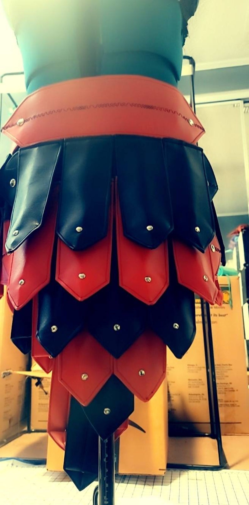 May include: A red and black leather skirt with a layered design. The skirt features alternating red and black panels, each secured with small silver rivets. The waist band is red and the skirt is displayed on a mannequin.