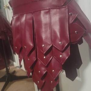 May include: A burgundy leather skirt with a layered design, featuring riveted accents. The skirt is displayed on a mannequin, showcasing its unique construction and style. The garment appears to be handmade.