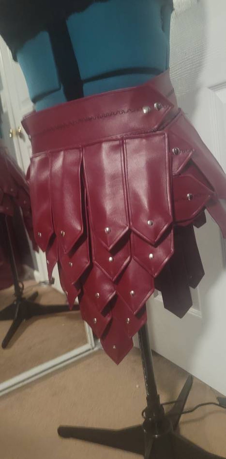 May include: A burgundy leather skirt with a layered design, featuring riveted accents. The skirt is displayed on a mannequin, showcasing its unique construction and style. The garment appears to be handmade.