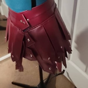 May include: A burgundy leather skirt with a layered, pleated design. The skirt is studded with silver rivets and is displayed on a mannequin. The garment is part of a fashion or costume collection.