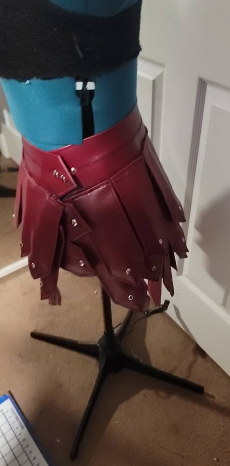 May include: A burgundy leather skirt with a layered, pleated design. The skirt is studded with silver rivets and is displayed on a mannequin. The garment is part of a fashion or costume collection.