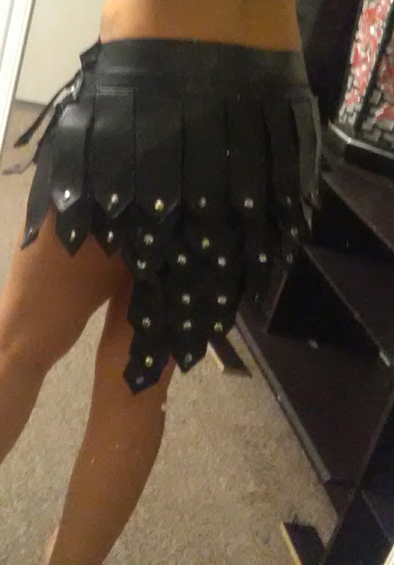 May include: Black leather skirt with a layered, segmented design. The skirt features small, silver-colored studs and a unique, edgy aesthetic. The skirt is a mini-skirt style.