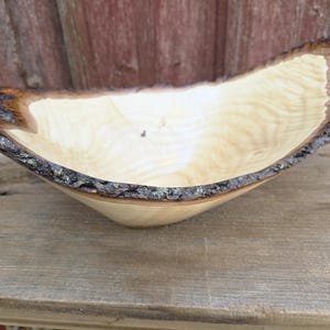 May include: A handcrafted wooden bowl with a natural edge. The bowl is light-colored with a smooth interior and a dark, textured bark edge. The bowl is displayed on a wooden surface.