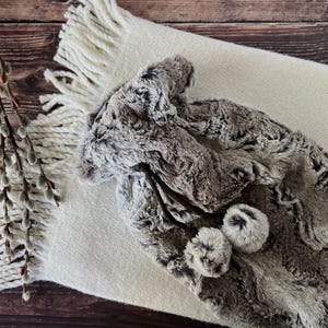 May include: A hot water bottle cover made of faux fur in shades of brown, grey, and white. The cover features a drawstring closure with two matching faux fur pom-poms attached to the ends of the dark brown cords, providing a soft texture.