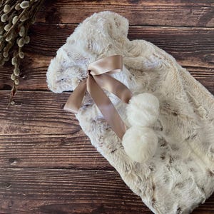 May include: A cream-coloured faux fur hot water bottle cover featuring a soft, plush texture. The cover is adorned with a smooth, light brown satin ribbon tied in a bow, accented by two fluffy, round cream-coloured pom-poms hanging from the ribbon.