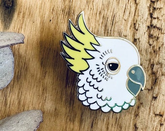 May include: White and yellow enamel pin of a cockatoo's head with a green beak. The pin is on a wooden surface.