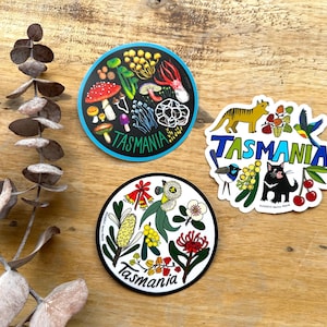 Könnte beinhalten: Drei runde Aufkleber mit bunten Illustrationen von tasmanischer Flora und Fauna. Der erste Aufkleber zeigt Pilze, Blumen und ein Blatt mit dem Wort "TASMANIA" in einem blauen Kreis. Der zweite Aufkleber zeigt einen Tasmanischen Teufel, einen Vogel und eine Katze mit dem Wort "TASMANIA" in einem blauen Kreis. Der dritte Aufkleber zeigt eine Vielzahl von Blumen und Blättern mit dem Wort "TASMANIA" in einem schwarzen Kreis.