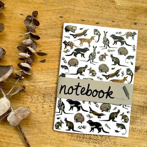May include: A white notebook with illustrations of Australian animals such as kangaroos and echidnas, and green leaves. The word "notebook" is printed on a grey band. A pencil illustration is on the right side of the band.