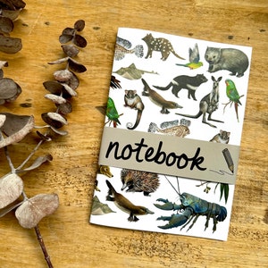 Könnte beinhalten: Ein weißes Notizbuch mit einer bunten Illustration australischer Tiere, darunter ein Schnabeltier, ein Ameisenigel, ein Wombat und ein Krebs. Das Notizbuch hat ein braunes Papierband mit dem Wort "notebook" in Schwarz.