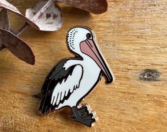 May include: A gold-tone enamel pin depicting a white pelican with black and grey markings. The pelican has a pink beak and is standing on a wooden surface.