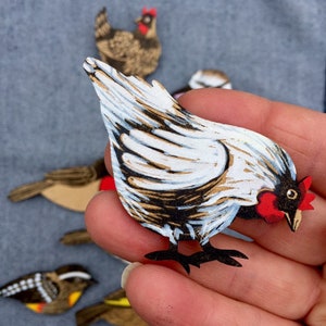 May include: A white and brown paper cut-out of a chicken with a red comb and wattle. The chicken is facing to the right and has its wings spread out. The chicken is being held in a person's hand.