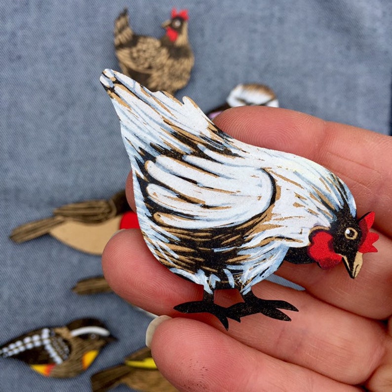 May include: A white and brown paper cut-out of a chicken with a red comb and wattle. The chicken is facing to the right and has its wings spread out. The chicken is being held in a person's hand.