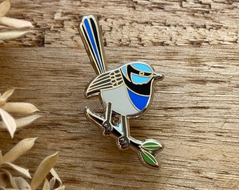 May include: A detailed enamel pin of a blue wren bird perched on a branch. The bird features a blue head, back, and tail, with a white chest and gray wings. The pin has a silver-colored metal outline and is set against a wooden background.