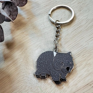 Op de afbeelding: Een grijze wombat sleutelhanger met een zilveren ketting en sleutelring. De wombat heeft zwarte accenten op zijn poten en neus, en witte highlights op zijn hoofd. Een schattig accessoire.
