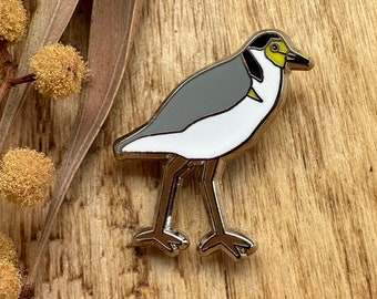 May include: A silver enamel pin featuring a cartoon illustration of a grey and white bird with a yellow head and black eyes. The bird is standing on two legs with its wings folded.