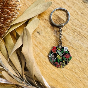 May include: A silver keychain with a round black enamel charm featuring a colorful illustration of Australian native flora and fauna. The charm includes a red flower, yellow wattle, a white flower, and a green bird.