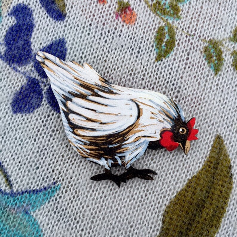 May include: A white and black painted wooden chicken brooch with a red comb. The brooch is on a white knitted fabric background.
