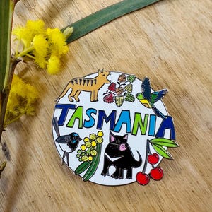 Puede incluir: Pin redondo de esmalte con la palabra "TASMANIA" en letras azules y verdes. El diseño incluye un tigre, pájaros, un demonio de Tasmania, cerezas y flora nativa, todo sobre un fondo blanco.