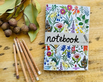 May include: A white notebook with a floral design featuring a variety of flowers and leaves in shades of pink, purple, yellow, and green. The notebook has the word "notebook" printed in black on a white band across the front.