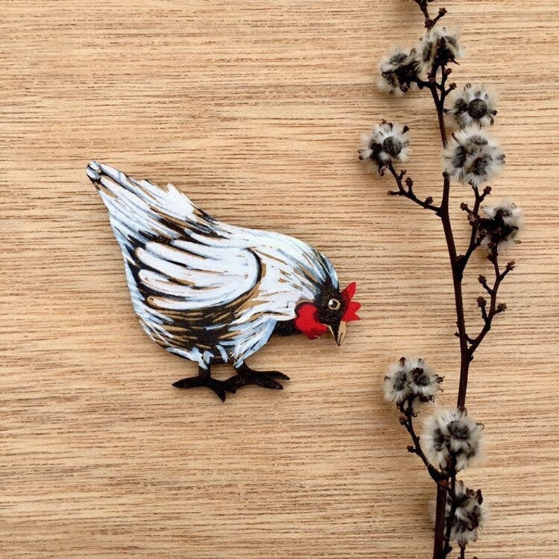 May include: A white wooden chicken figurine with black markings and a red comb. The chicken is looking down and has its wings spread out. The figurine is painted in a realistic style and is set against a wooden background.