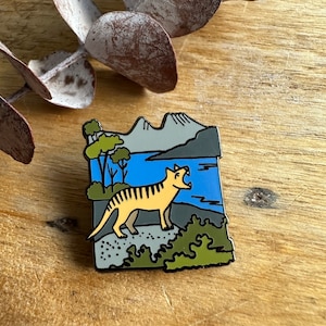 Pode incluir: Um broche de esmalte amarelo e preto com um tigre da Tasmânia a uivar em frente a uma paisagem montanhosa. O broche está sobre um fundo de madeira.