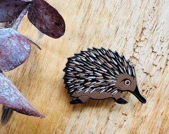 May include: A brown and white enamel pin of an echidna, a spiny anteater native to Australia and New Guinea. The pin is on a wooden surface.