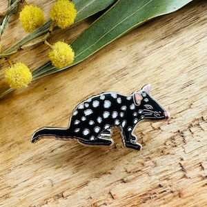 Pode incluir: Um broche de esmalte preto e branco com um quol manchado, um pequeno marsupial nativo da Austrália. O quol é retratado em uma posição agachada com a cauda enrolada ao redor do seu corpo.