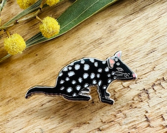 May include: A black and white enamel pin depicting a spotted quoll, a small marsupial native to Australia. The quoll is shown in a crouching position with its tail curled around its body.