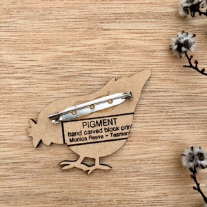 May include: A wooden bird brooch with a pin on the back. The brooch is hand carved and features the text "PIGMENT hand carved block print Monica Reeve - Tasmania".