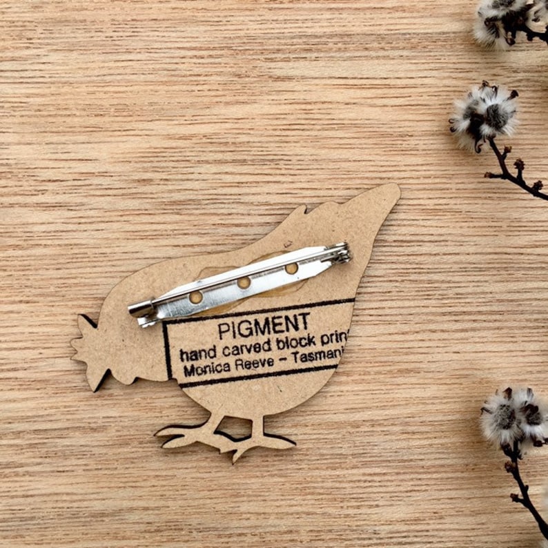 May include: A wooden bird brooch with a pin on the back. The brooch is hand carved and features the text "PIGMENT hand carved block print Monica Reeve - Tasmania".