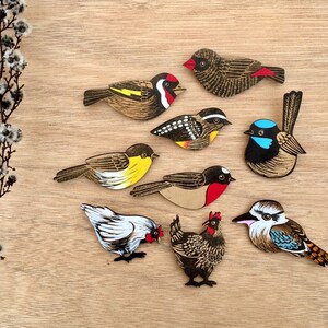 May include: A set of nine wooden bird cutouts, each painted with intricate details and vibrant colors. The birds include a robin, a bluebird, a wren, a sparrow, a finch, a chickadee, a cardinal, a hummingbird, and a woodpecker.