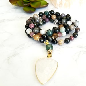 May include: A multi-strand mala necklace with black, brown, green, and white beads. The necklace has a white stone pendant with a gold trim.