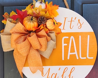 May include: A round wooden door hanger with the text "It's Fall Ya'll" in white lettering on an orange and white background. The hanger is decorated with a burlap bow, fall leaves, and small pumpkins.