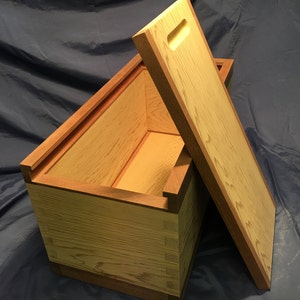 May include: A wooden box with a lid ajar, showcasing a natural wood grain. The box features dovetail joints and a contrasting darker wood trim. The lid has a handle cutout. The box is likely for storage or decorative purposes.