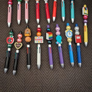 May include: A collection of seventeen multi-color ink pens featuring decorative beaded toppers with various school-themed charms including apples, books, clocks, and the word TEACHER. Each pen has a multi-colored barrel and a unique arrangement of beads.