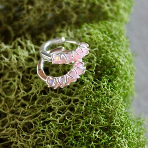 May include: A pair of silver hoop earrings with pink gemstones, resting on a bed of vibrant green moss. The earrings are small and delicate, with the gemstones adding a touch of color and sparkle to the design.
