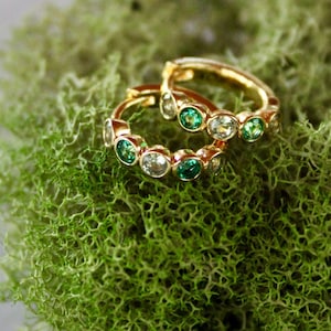 May include: Two gold hoop earrings, each adorned with a series of green and clear gemstones. The earrings rest on a bed of vibrant green moss, set against a gray background. The jewelry is a delicate and elegant accessory.