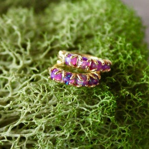 Può includere: Un paio di orecchini a cerchio color oro con pietre preziose rosa e viola, appoggiati su un letto di muschio verde vibrante. Gli orecchini sono piccoli e delicati, che mostrano un design colorato.